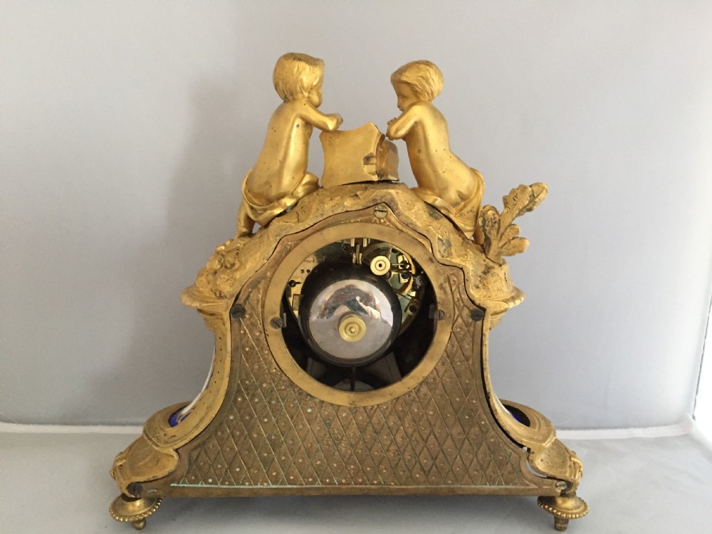An antique French gilt bronze ormolu mantel clock featuring sculpted cherubs on top and hand-painted blue porcelain panels around a Roman numeral dial. View X
