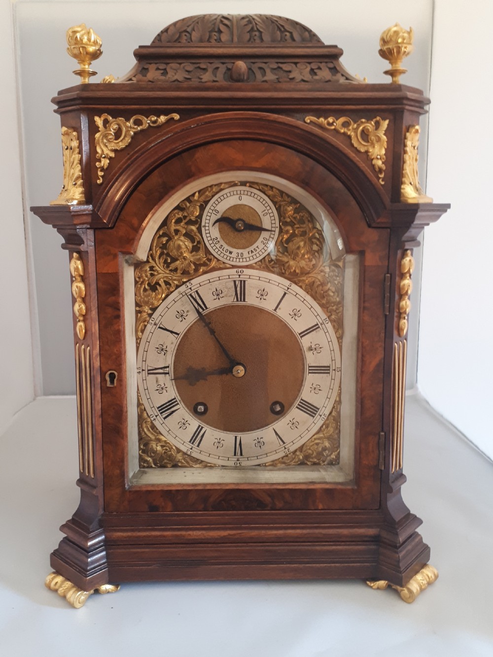 An arched burr walnut bracket clock with ornate gilt brass mounts, finials, and an intricate brass dial displaying Roman numerals.
