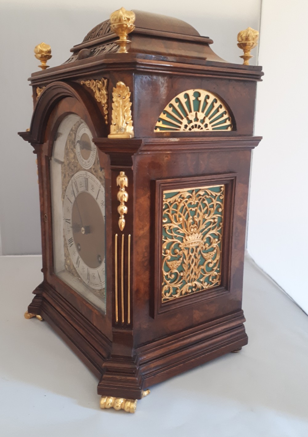 An arched burr walnut bracket clock with ornate gilt brass mounts, finials, and an intricate brass dial displaying Roman numerals. View VI