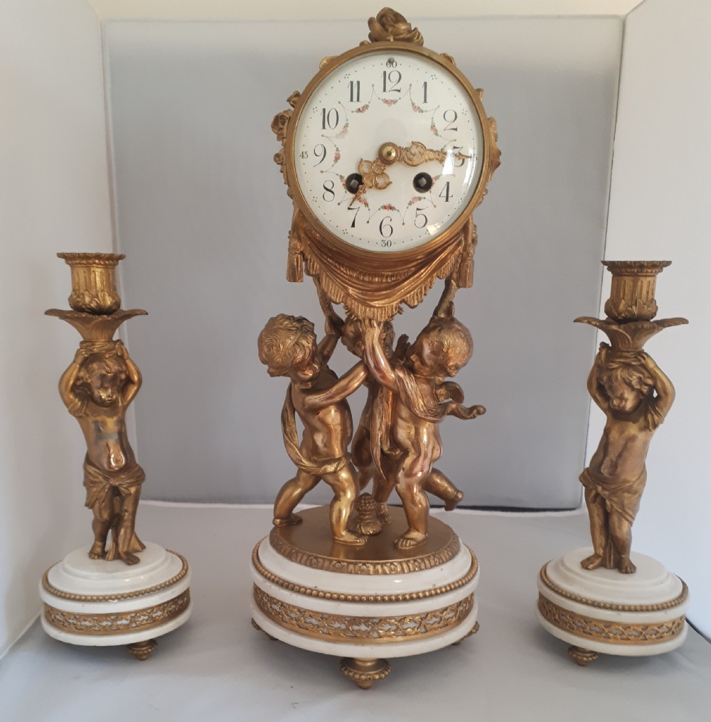 A three-piece French gilt metal and white marble clock garniture featuring cherub figures supporting a central clock drum alongside two matching single-light candelabras.