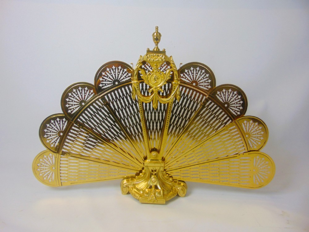 An antique French gilt bronze folding fan fire screen featuring intricate pierced lattice panels, a central crest with a relief medallion, and a weighted cast scroll base.