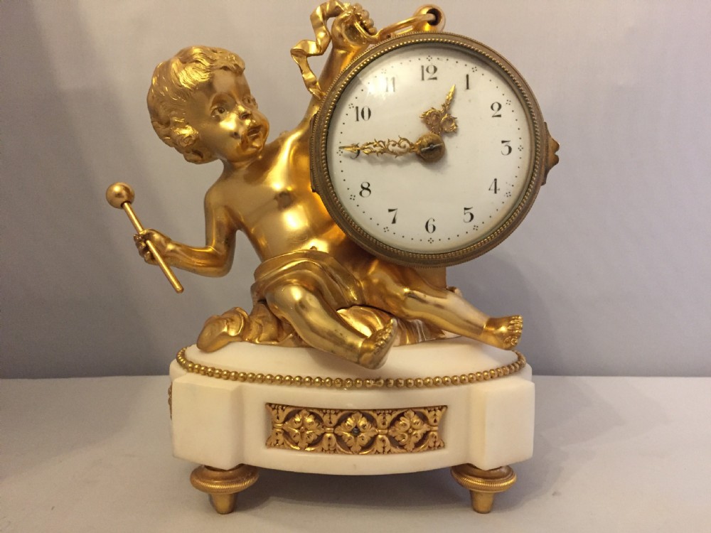 A gilt bronze figure of a cherub reclining on a white marble base while supporting a circular white enamel clock face.