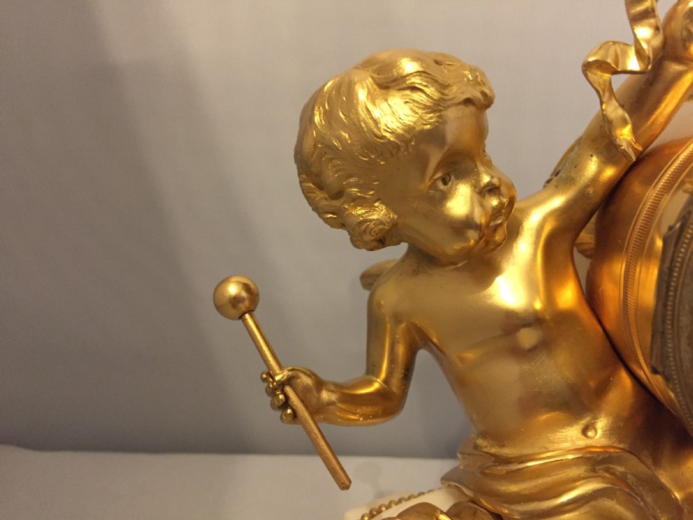A gilt bronze figure of a cherub reclining on a white marble base while supporting a circular white enamel clock face. View II