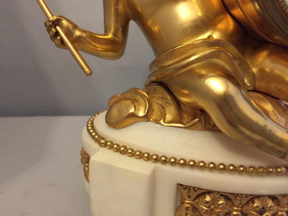 A gilt bronze figure of a cherub reclining on a white marble base while supporting a circular white enamel clock face. View III