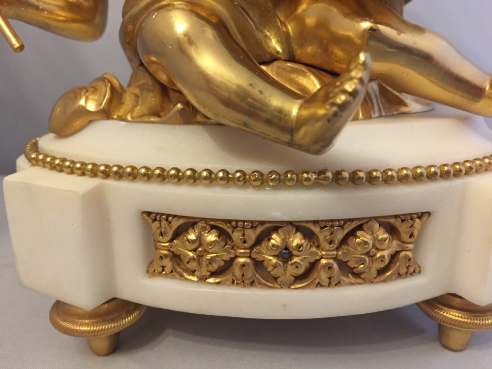 A gilt bronze figure of a cherub reclining on a white marble base while supporting a circular white enamel clock face. View V