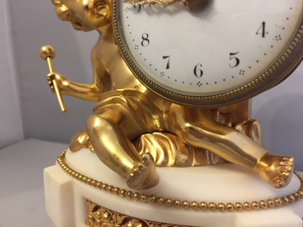 A gilt bronze figure of a cherub reclining on a white marble base while supporting a circular white enamel clock face. View VI