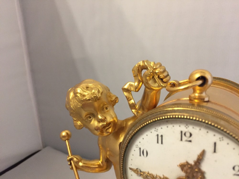 A gilt bronze figure of a cherub reclining on a white marble base while supporting a circular white enamel clock face. View VII
