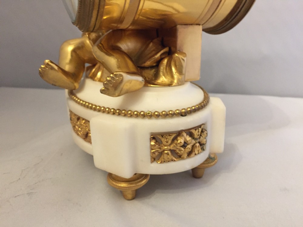 A gilt bronze figure of a cherub reclining on a white marble base while supporting a circular white enamel clock face. View VIII
