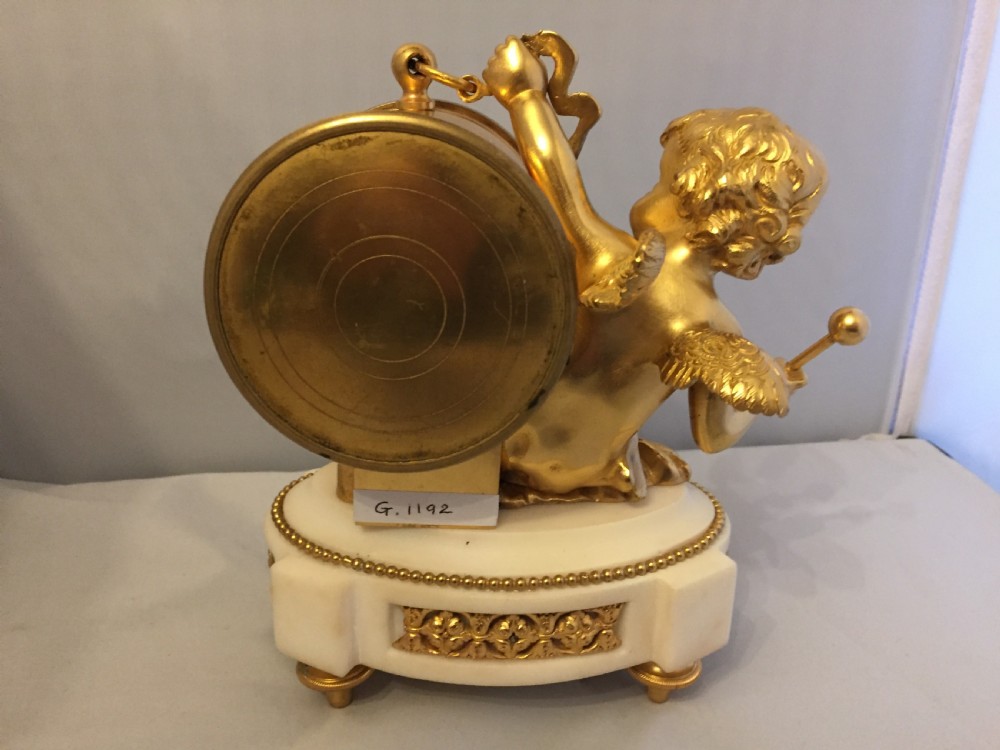 A gilt bronze figure of a cherub reclining on a white marble base while supporting a circular white enamel clock face. View X