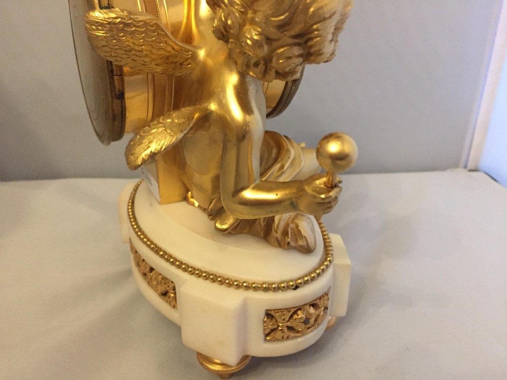 A gilt bronze figure of a cherub reclining on a white marble base while supporting a circular white enamel clock face. View XII