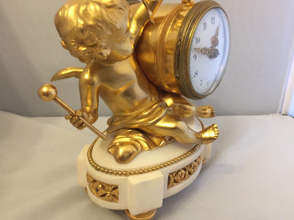 A gilt bronze figure of a cherub reclining on a white marble base while supporting a circular white enamel clock face. View XIII