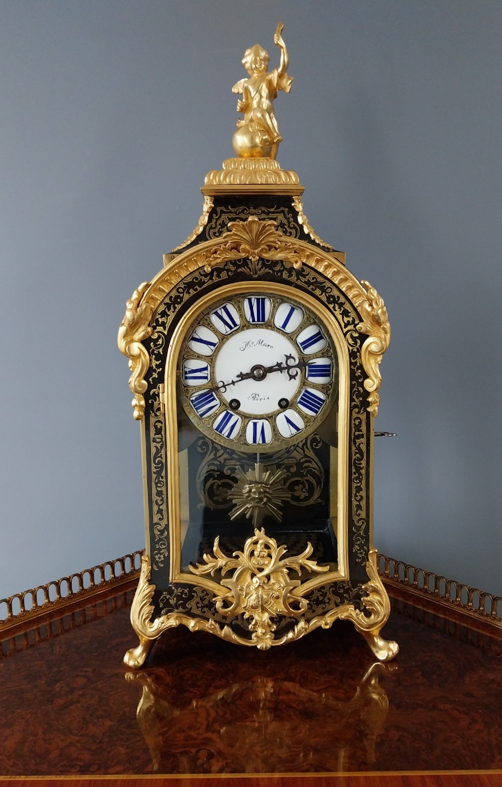 A French Rococo Revival Boulle mantel clock featuring brass marquetry inlay, gilt bronze mounts, an enamel cartouche dial, and a cherub finial.