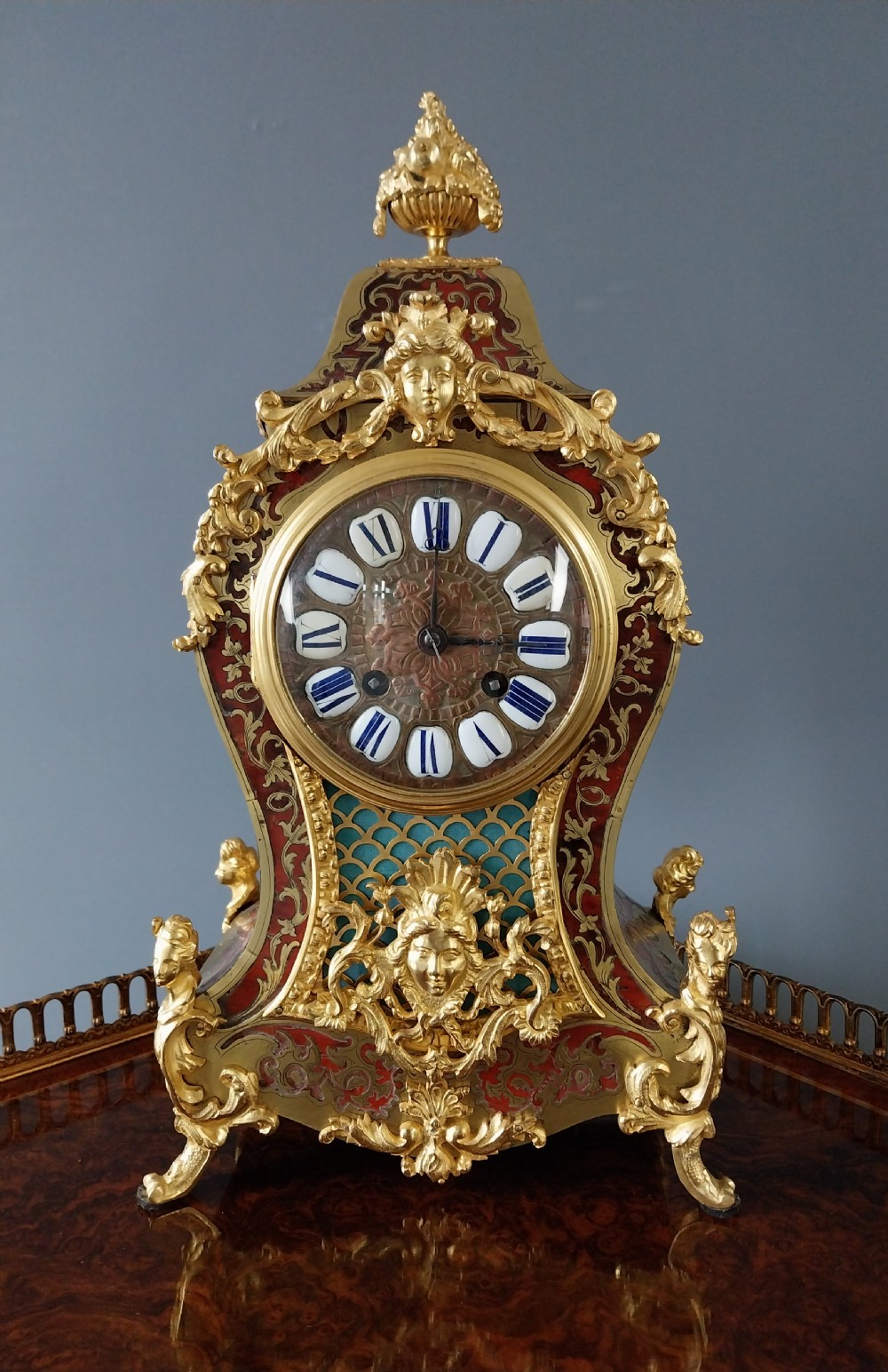 An ornate French Rococo style mantel clock featuring red tortoiseshell Boulle marquetry, decorative gilt bronze mounts, and blue enamel Roman numeral cartouches.
