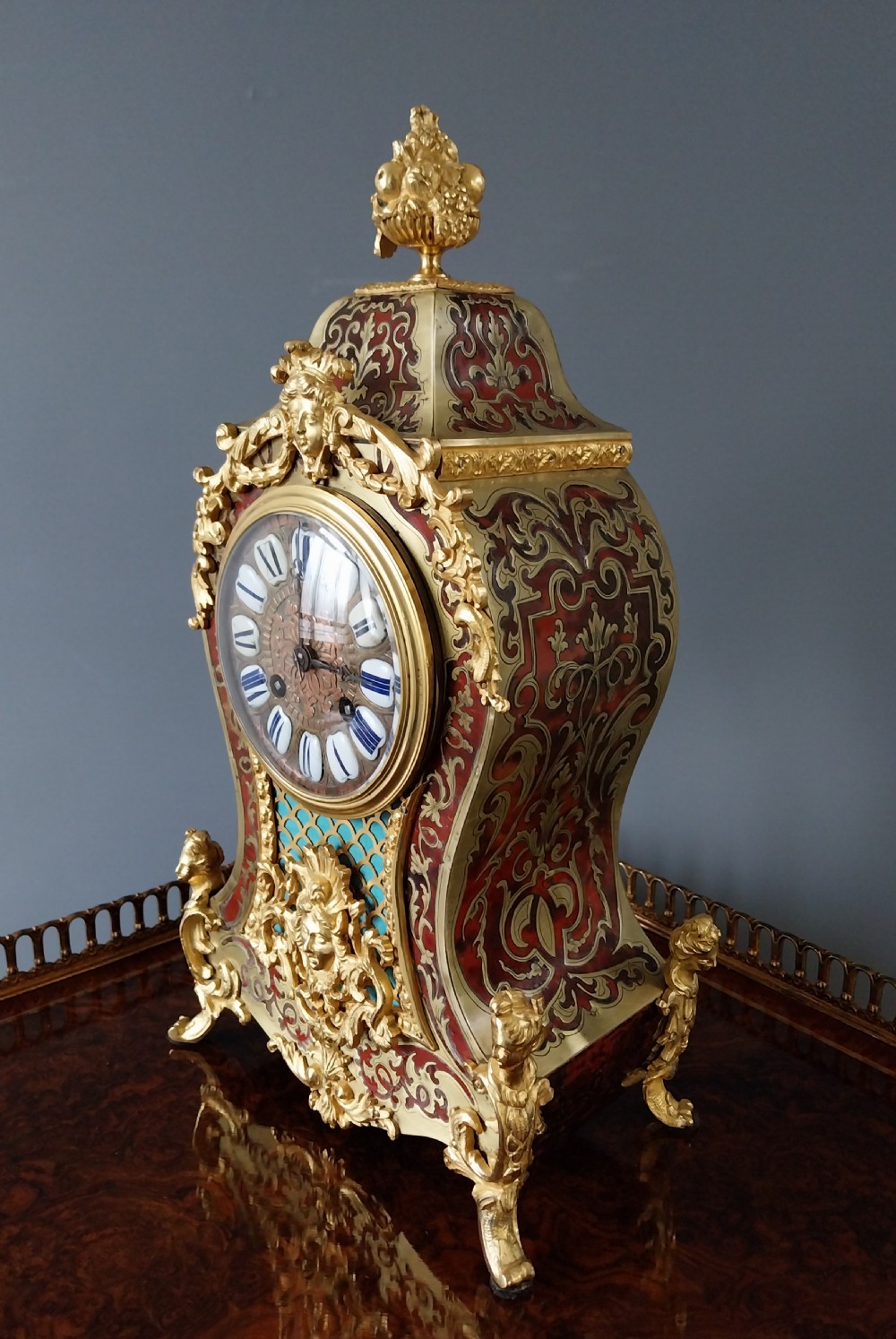 An ornate French Rococo style mantel clock featuring red tortoiseshell Boulle marquetry, decorative gilt bronze mounts, and blue enamel Roman numeral cartouches. View II