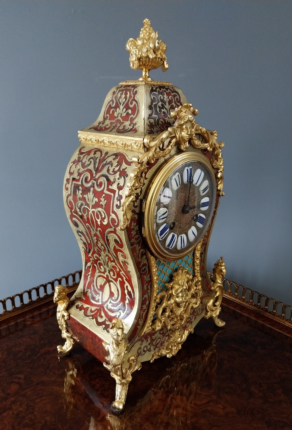 An ornate French Rococo style mantel clock featuring red tortoiseshell Boulle marquetry, decorative gilt bronze mounts, and blue enamel Roman numeral cartouches. View III