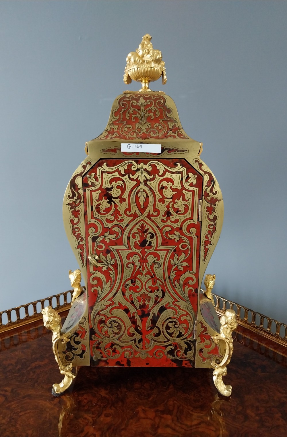 An ornate French Rococo style mantel clock featuring red tortoiseshell Boulle marquetry, decorative gilt bronze mounts, and blue enamel Roman numeral cartouches. View V