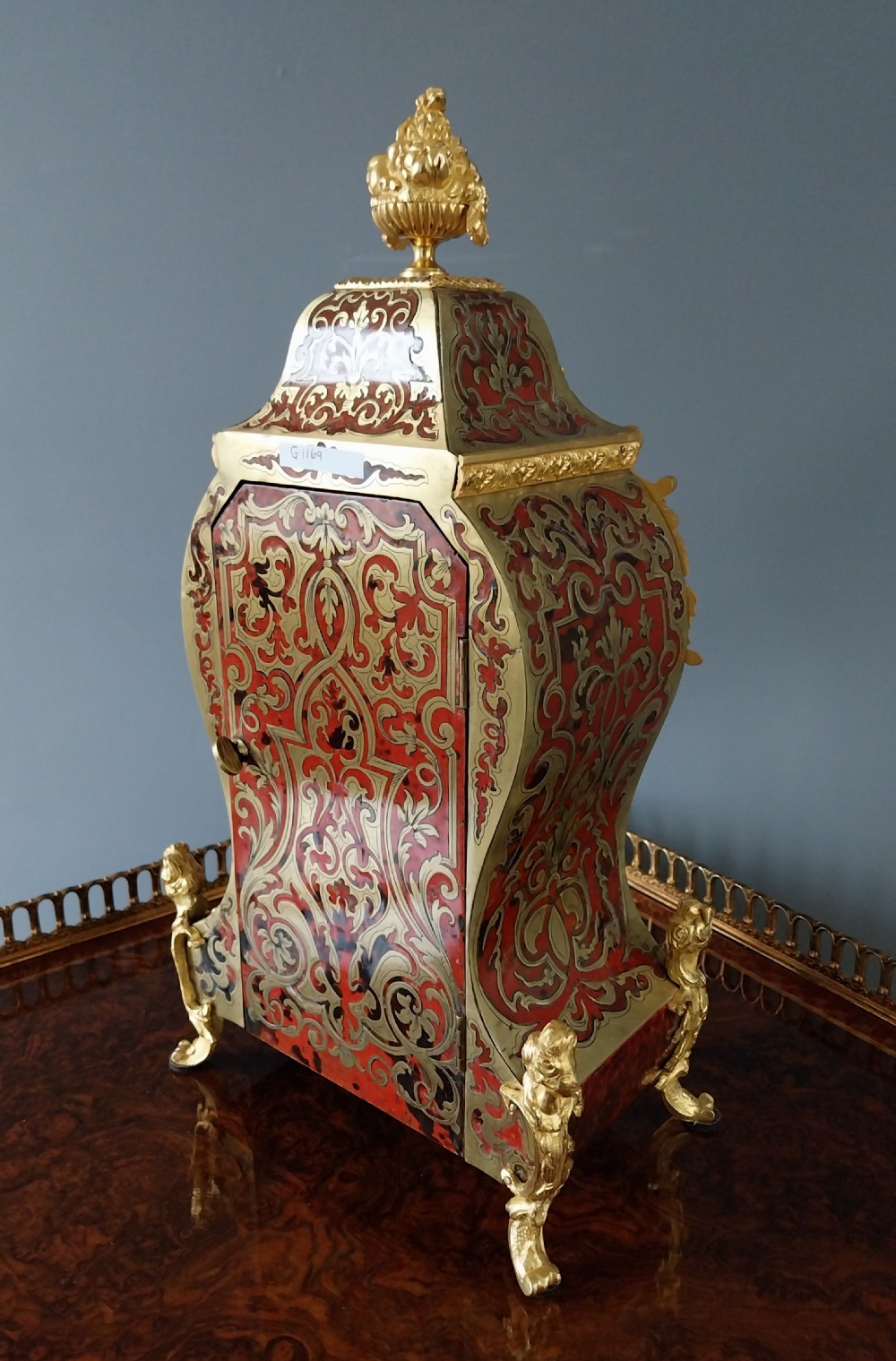 An ornate French Rococo style mantel clock featuring red tortoiseshell Boulle marquetry, decorative gilt bronze mounts, and blue enamel Roman numeral cartouches. View VI