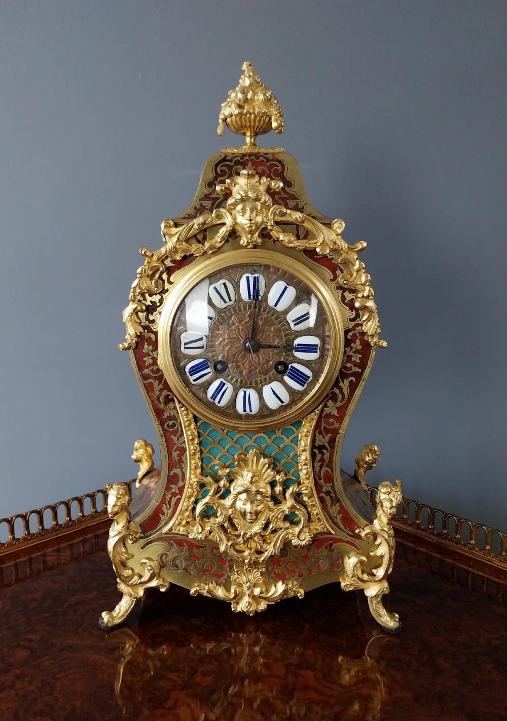 An ornate French Rococo style mantel clock featuring red tortoiseshell Boulle marquetry, decorative gilt bronze mounts, and blue enamel Roman numeral cartouches. View VII