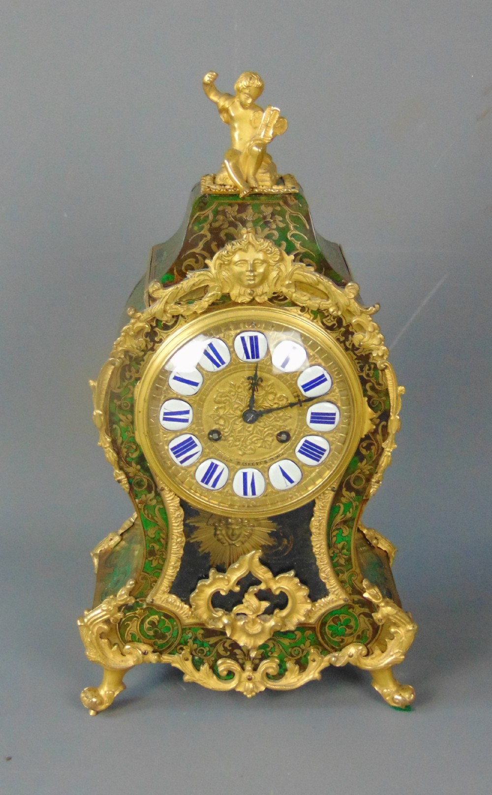 A French Rococo style green Boulle mantel clock featuring brass marquetry inlay, gilded ormolu mounts, a putto finial, and blue enamelled Roman numeral cartouches.