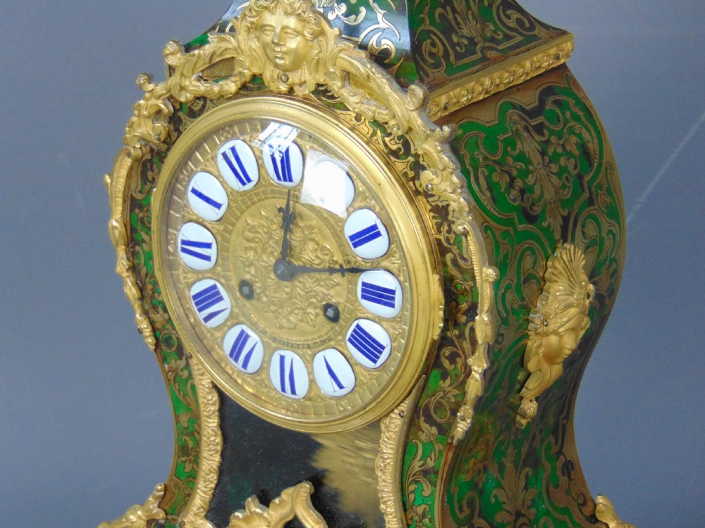 A French Rococo style green Boulle mantel clock featuring brass marquetry inlay, gilded ormolu mounts, a putto finial, and blue enamelled Roman numeral cartouches. View V