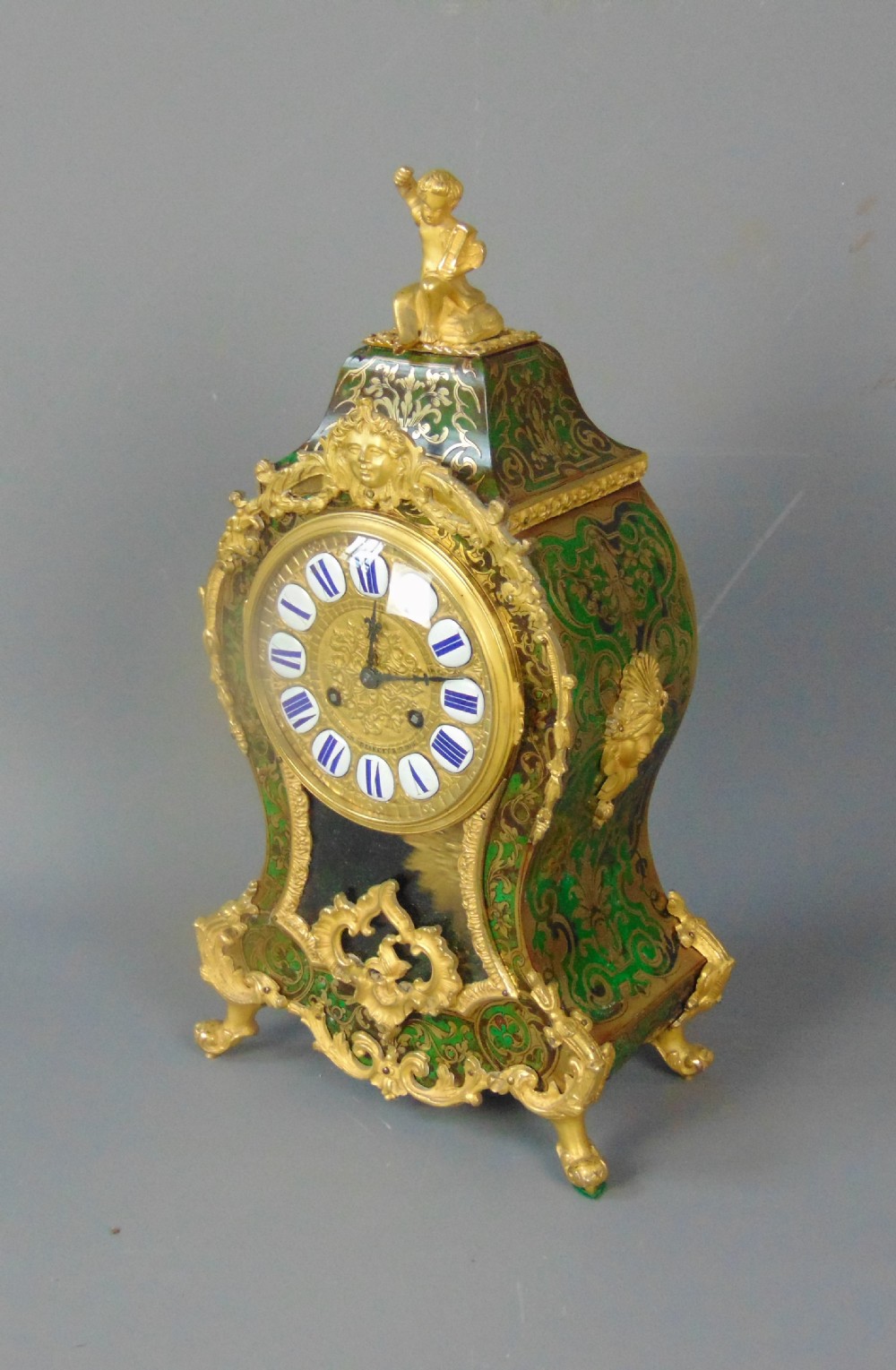A French Rococo style green Boulle mantel clock featuring brass marquetry inlay, gilded ormolu mounts, a putto finial, and blue enamelled Roman numeral cartouches. View VI