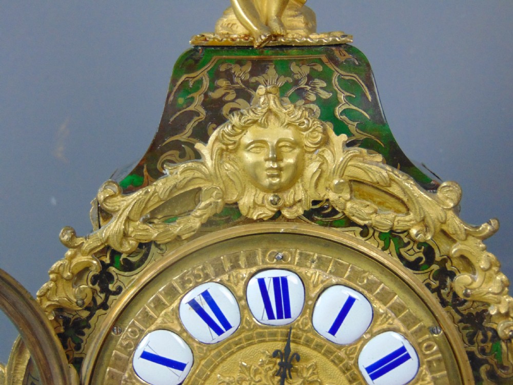 A French Rococo style green Boulle mantel clock featuring brass marquetry inlay, gilded ormolu mounts, a putto finial, and blue enamelled Roman numeral cartouches. View VIII