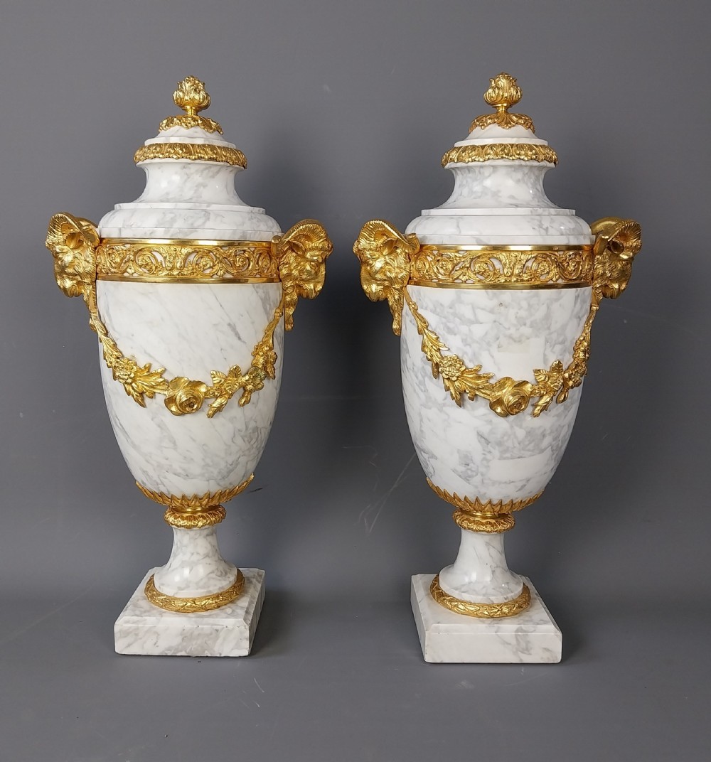A pair of white veined marble cassolette urns decorated with gilt bronze mounts, floral swags, ram head handles, and finial lids on square bases.