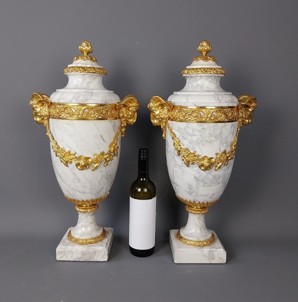 A pair of white veined marble cassolette urns decorated with gilt bronze mounts, floral swags, ram head handles, and finial lids on square bases. View II