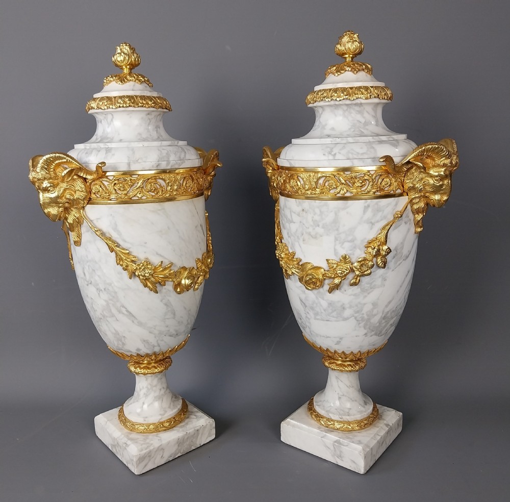 A pair of white veined marble cassolette urns decorated with gilt bronze mounts, floral swags, ram head handles, and finial lids on square bases. View III