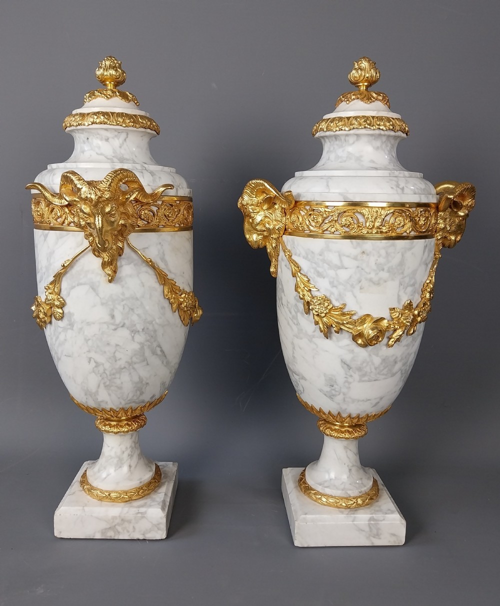 A pair of white veined marble cassolette urns decorated with gilt bronze mounts, floral swags, ram head handles, and finial lids on square bases. View V