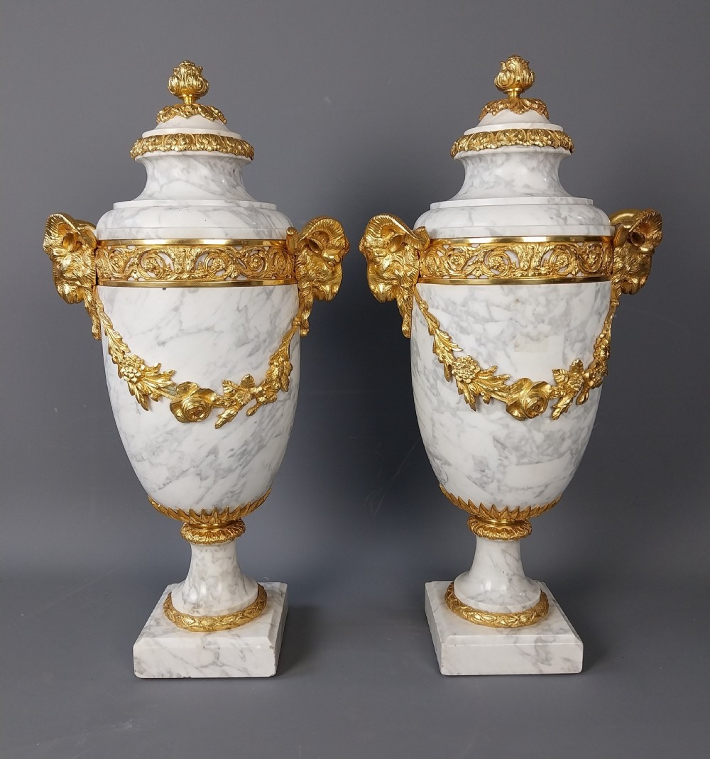 A pair of white veined marble cassolette urns decorated with gilt bronze mounts, floral swags, ram head handles, and finial lids on square bases. View VI