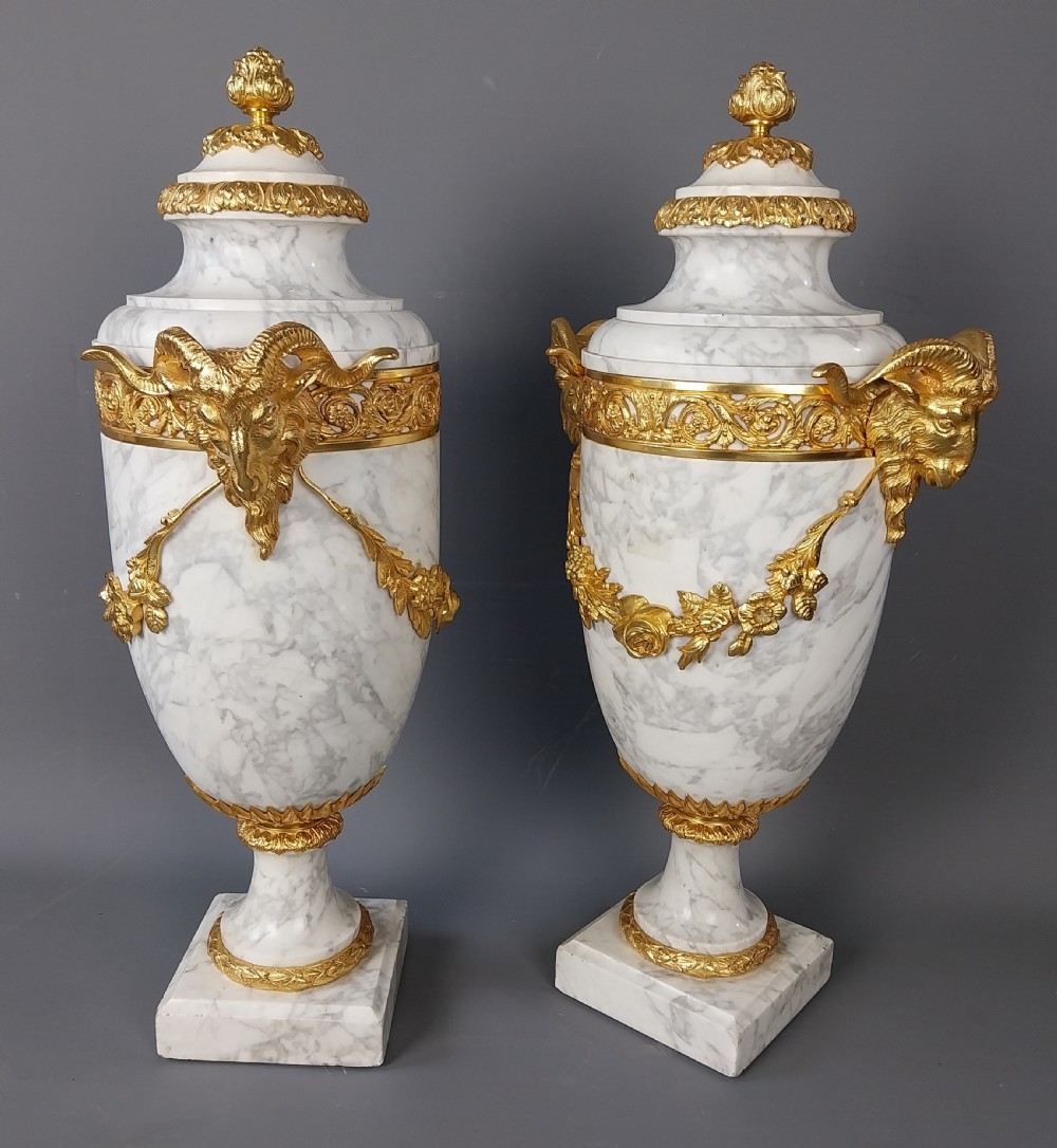 A pair of white veined marble cassolette urns decorated with gilt bronze mounts, floral swags, ram head handles, and finial lids on square bases. View VII