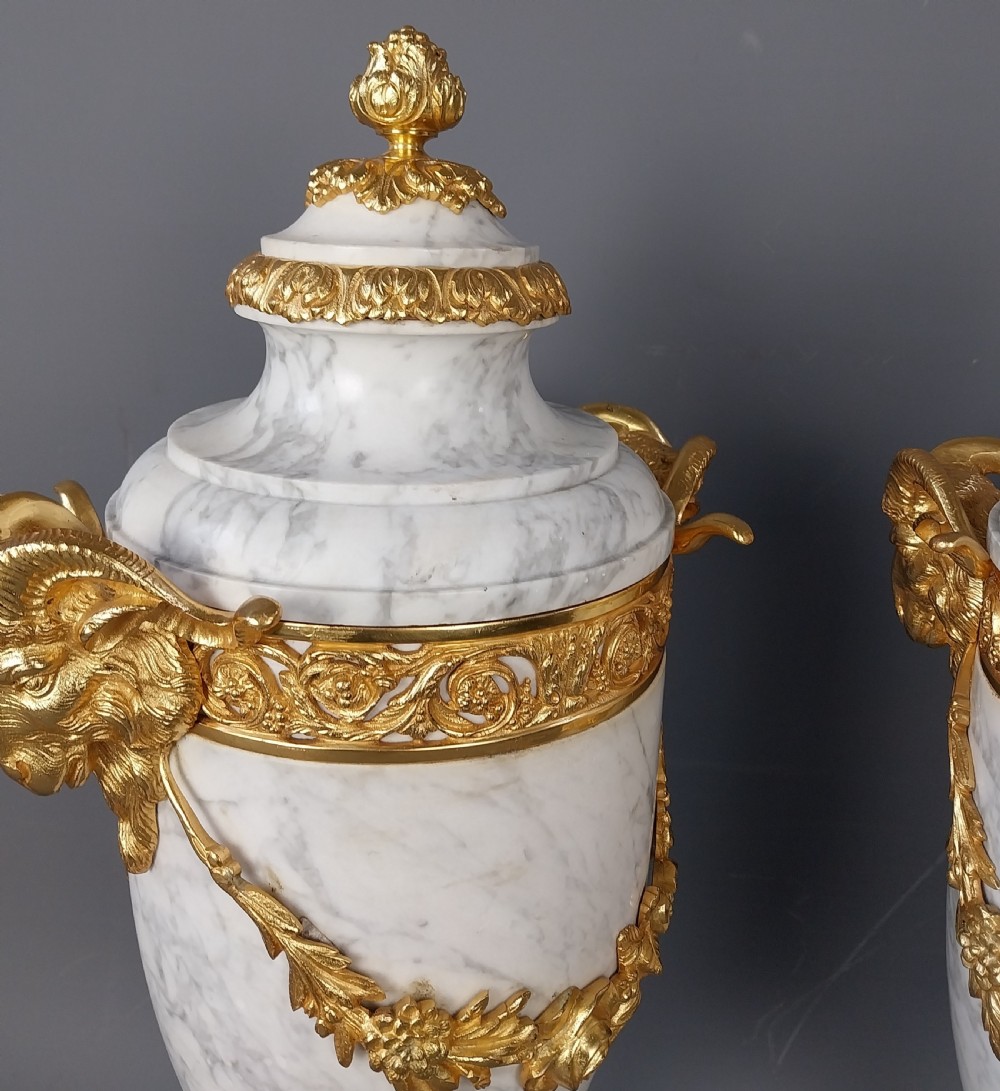 A pair of white veined marble cassolette urns decorated with gilt bronze mounts, floral swags, ram head handles, and finial lids on square bases. View VIII