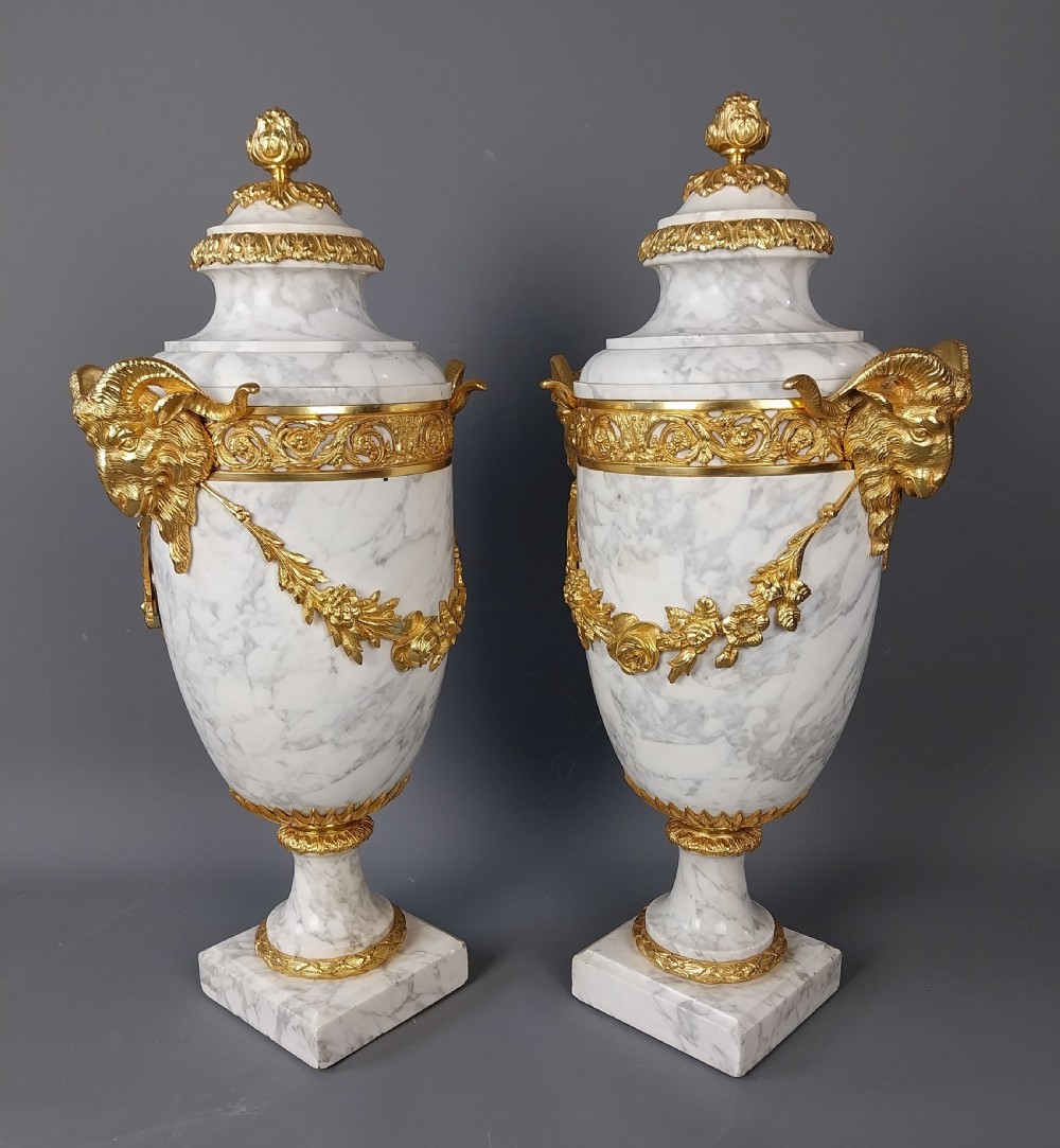 A pair of white veined marble cassolette urns decorated with gilt bronze mounts, floral swags, ram head handles, and finial lids on square bases. View IX