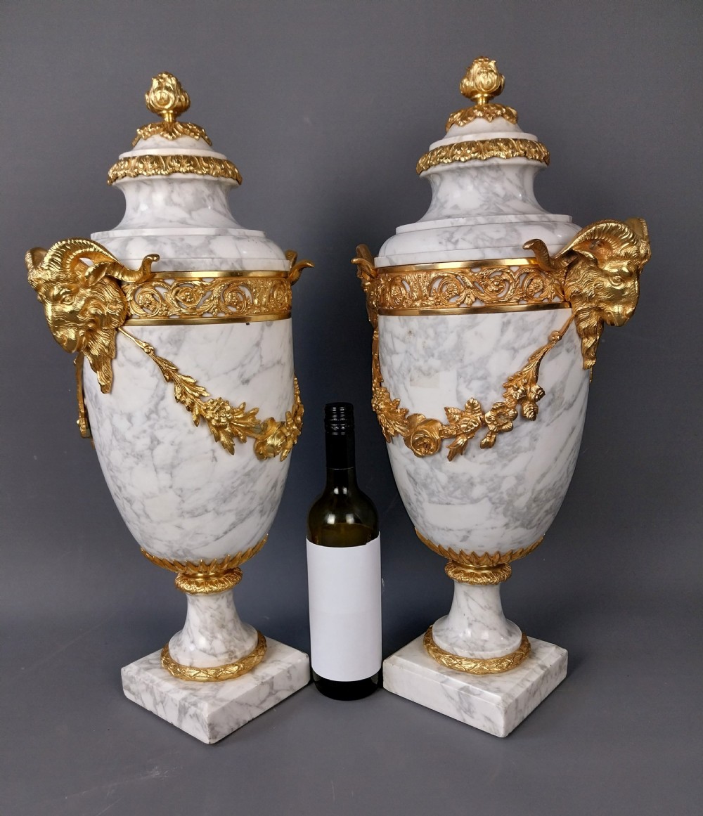 A pair of white veined marble cassolette urns decorated with gilt bronze mounts, floral swags, ram head handles, and finial lids on square bases. View X