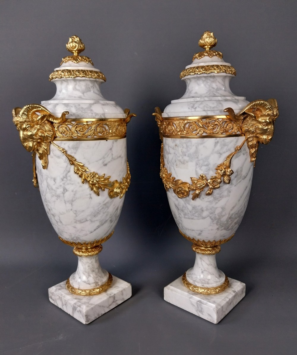 A pair of white veined marble cassolette urns decorated with gilt bronze mounts, floral swags, ram head handles, and finial lids on square bases. View XI