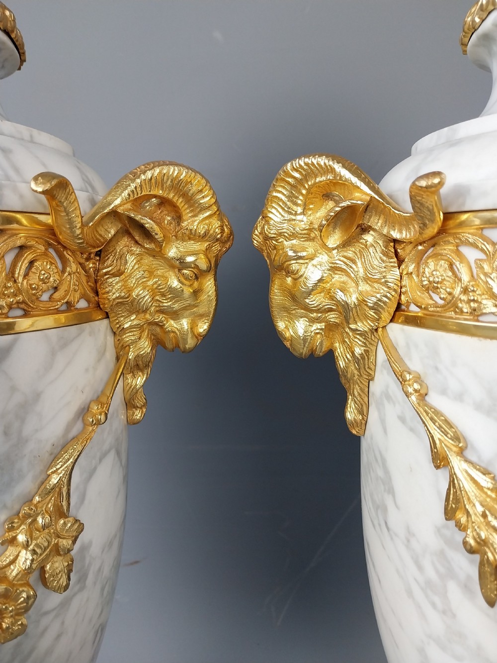 A pair of white veined marble cassolette urns decorated with gilt bronze mounts, floral swags, ram head handles, and finial lids on square bases. View XII