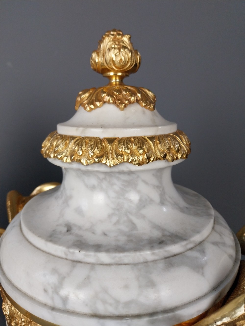 A pair of white veined marble cassolette urns decorated with gilt bronze mounts, floral swags, ram head handles, and finial lids on square bases. View XV