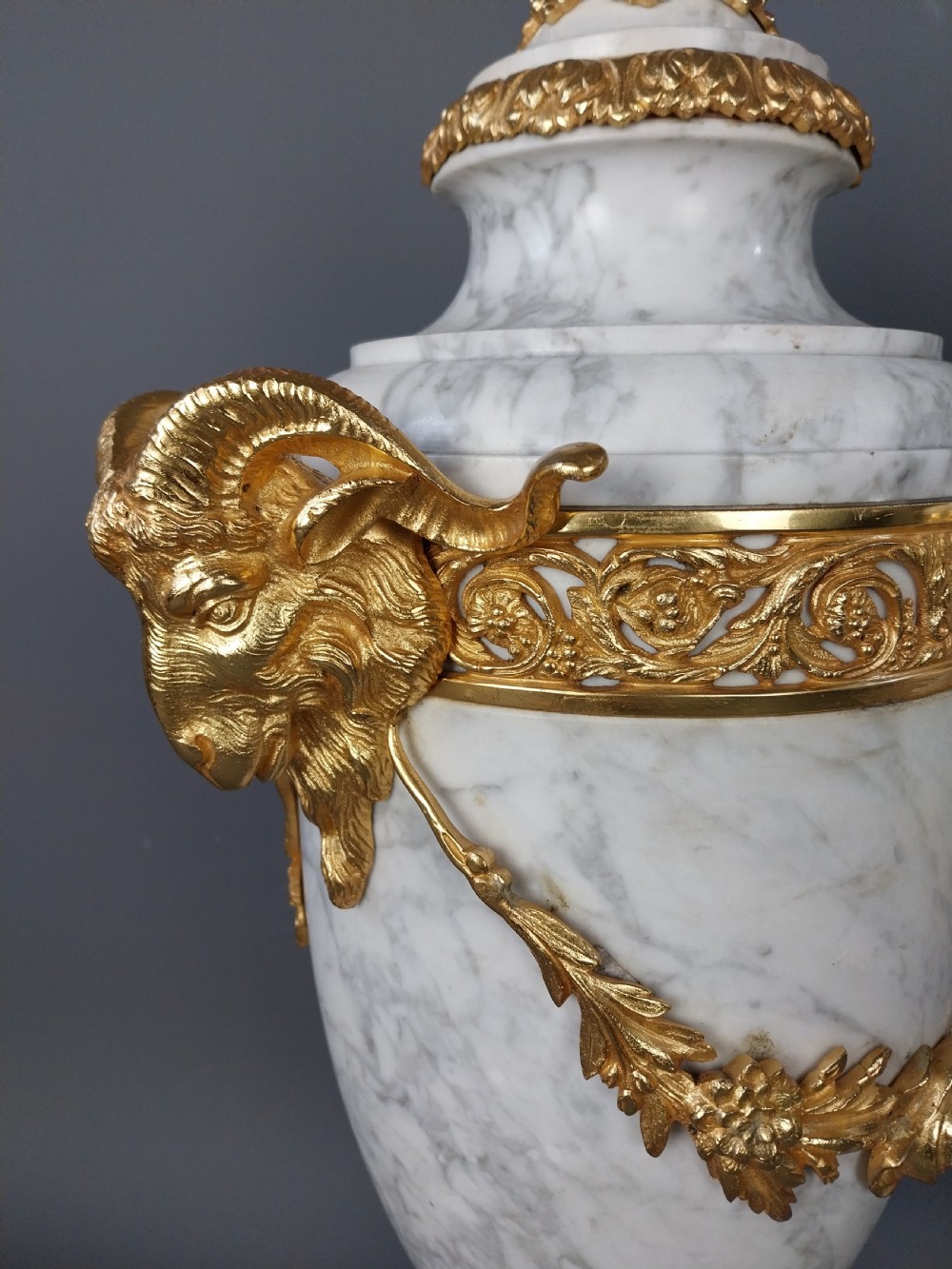 A pair of white veined marble cassolette urns decorated with gilt bronze mounts, floral swags, ram head handles, and finial lids on square bases. View XVI
