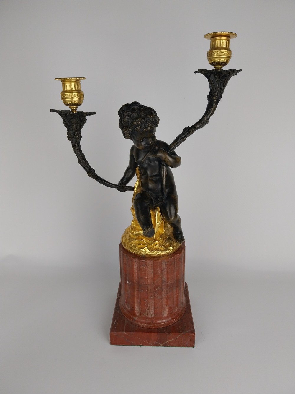 A patinated bronze cherub figure holding two scrolling branches with gilt sconces, set upon a round fluted red marble plinth.