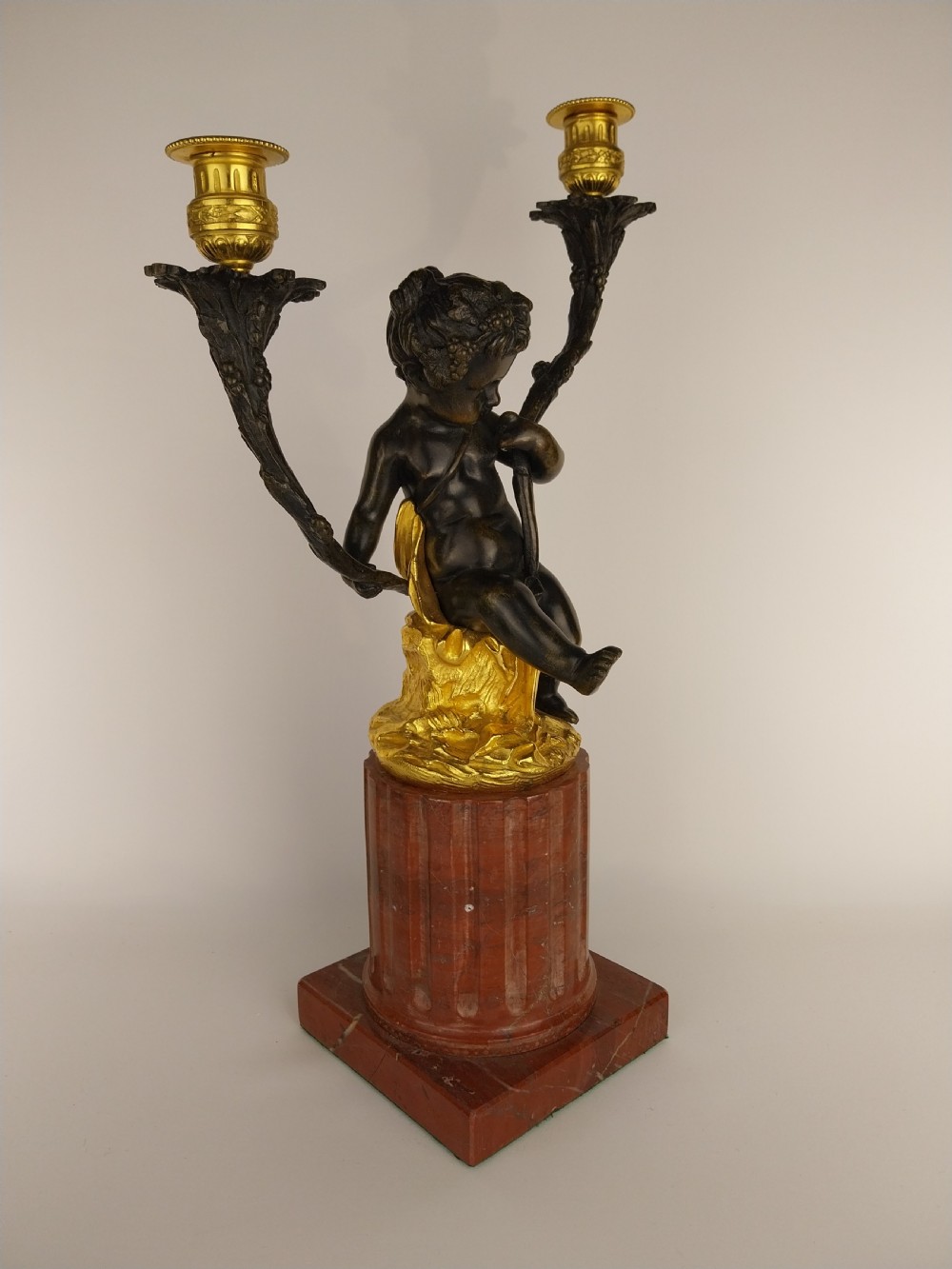 A patinated bronze cherub figure holding two scrolling branches with gilt sconces, set upon a round fluted red marble plinth. View II
