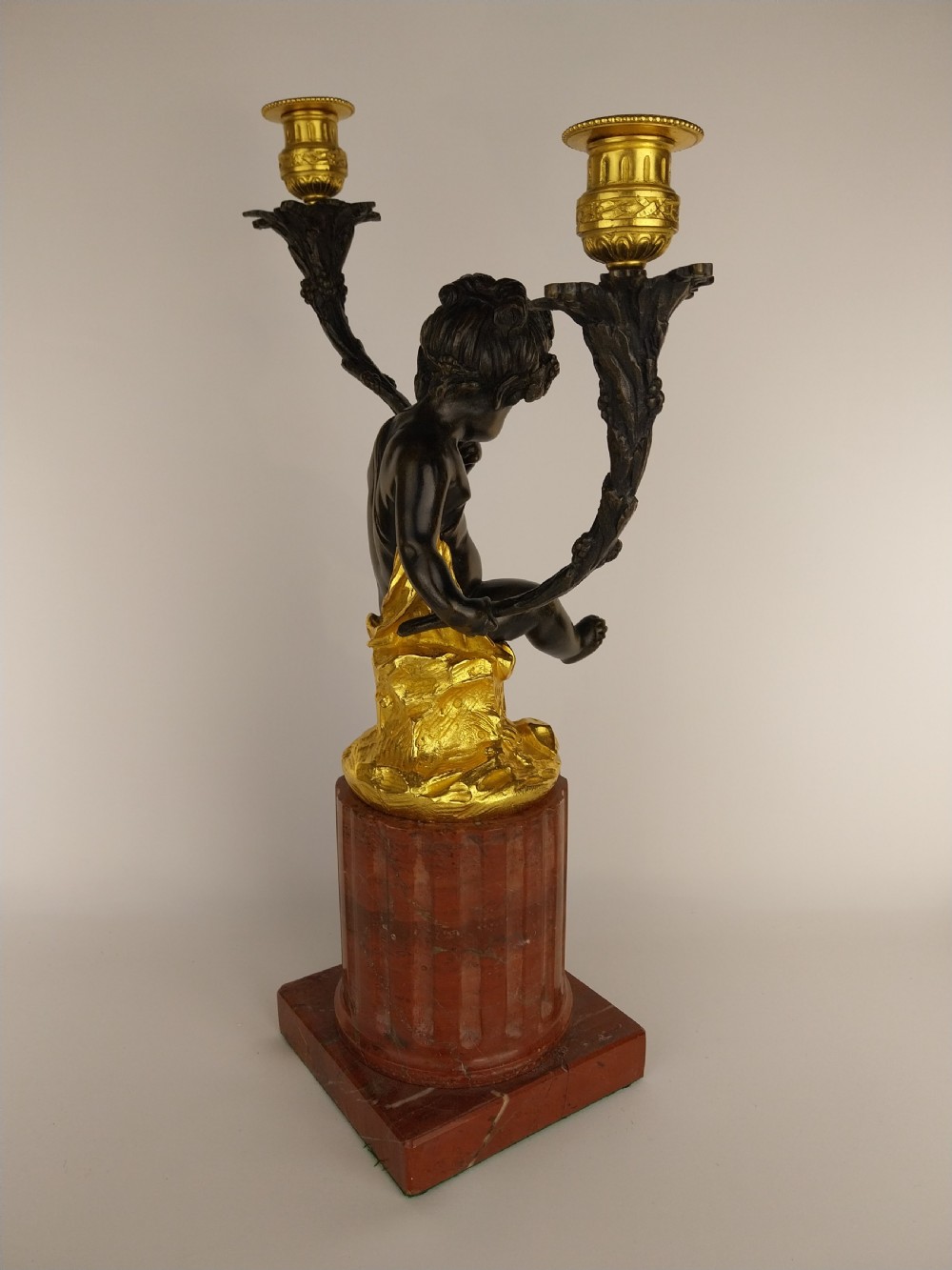 A patinated bronze cherub figure holding two scrolling branches with gilt sconces, set upon a round fluted red marble plinth. View III