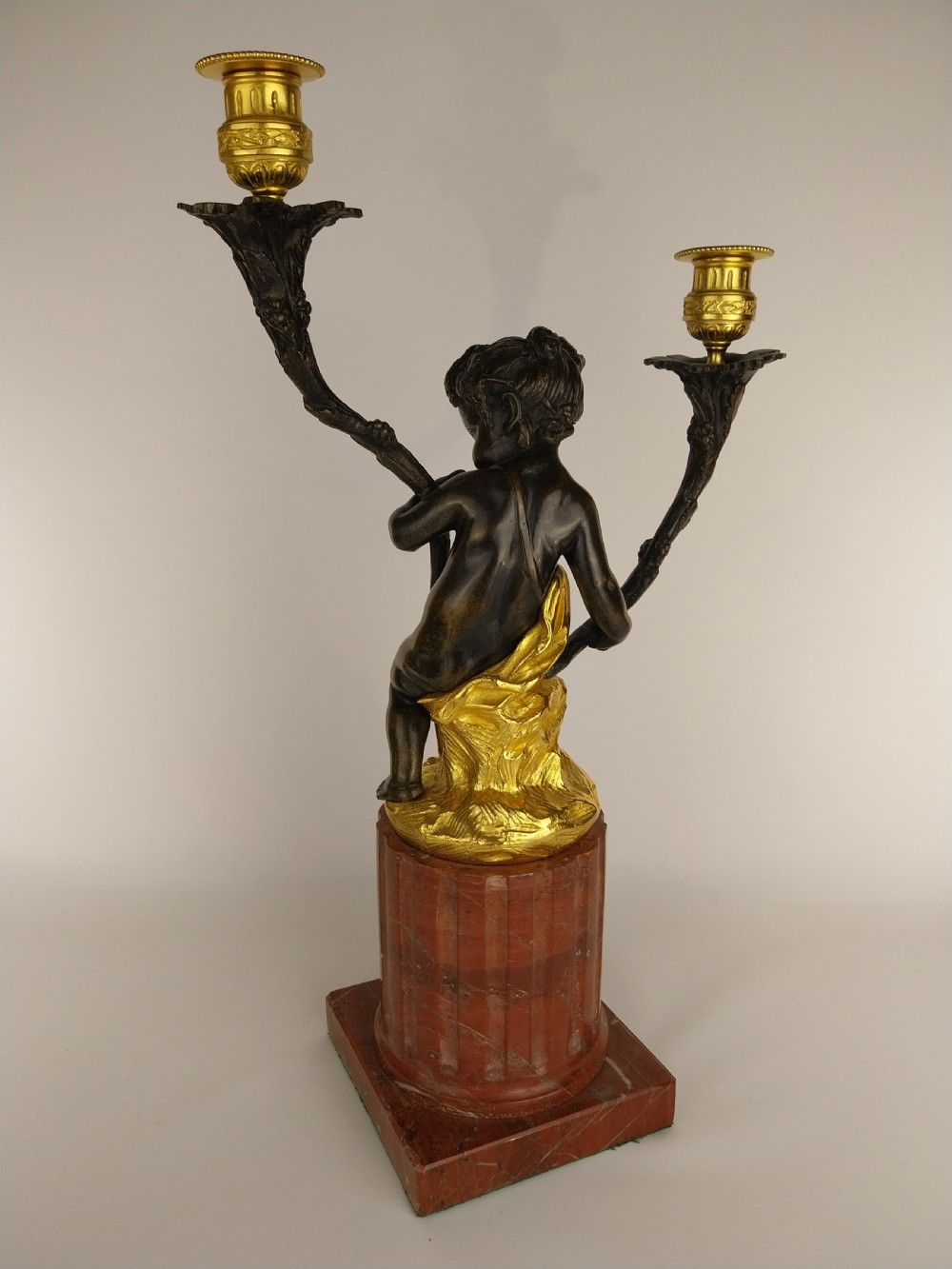 A patinated bronze cherub figure holding two scrolling branches with gilt sconces, set upon a round fluted red marble plinth. View IV