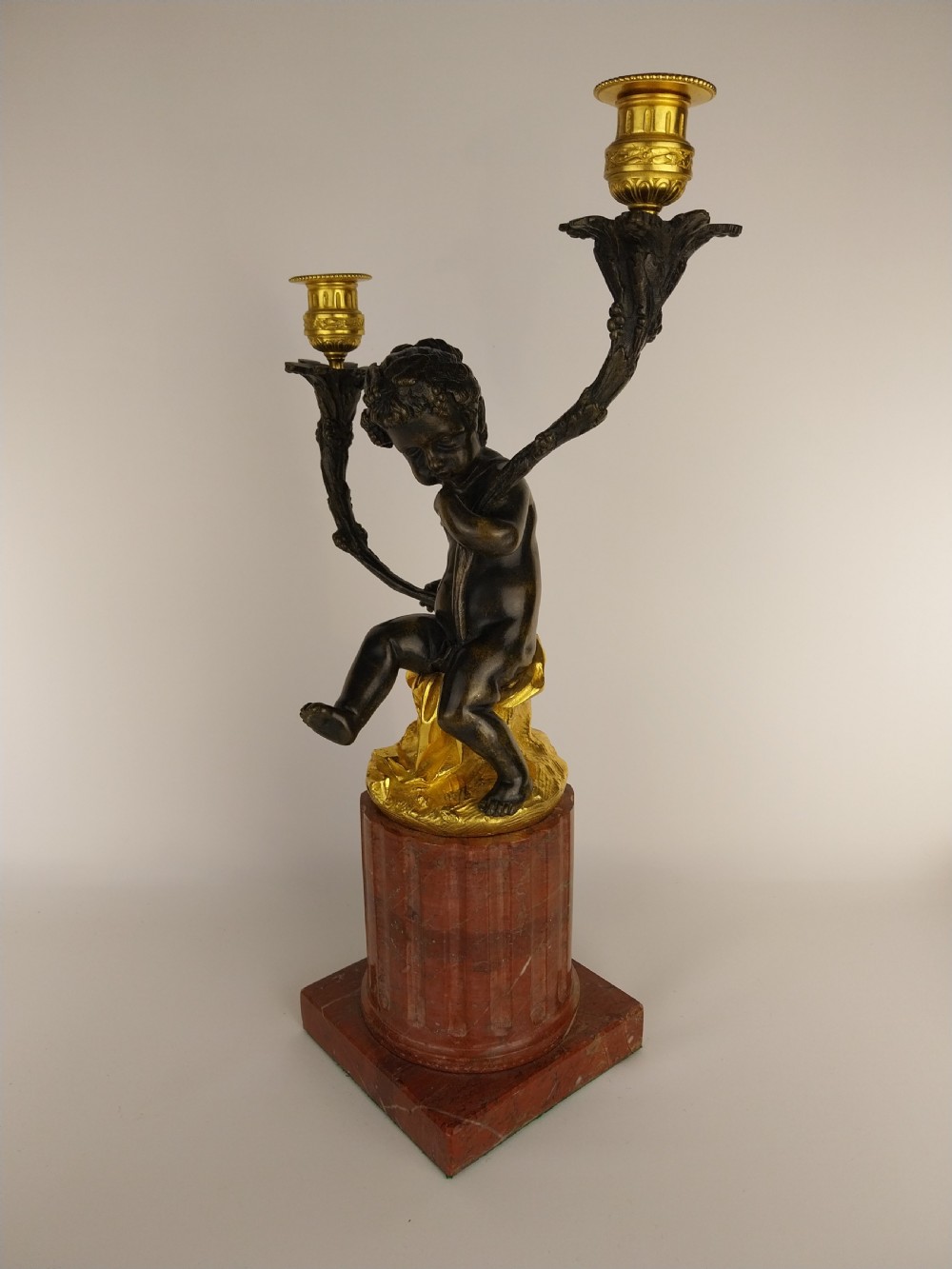 A patinated bronze cherub figure holding two scrolling branches with gilt sconces, set upon a round fluted red marble plinth. View VII