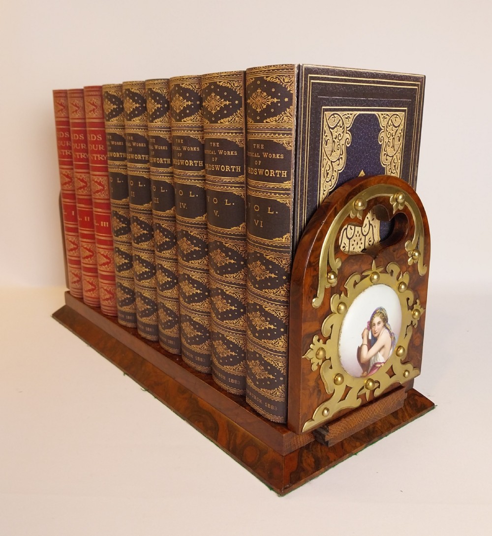 A Victorian burr walnut sliding book slide features pierced brass mounts, decorative studs, and a central painted porcelain plaque.
