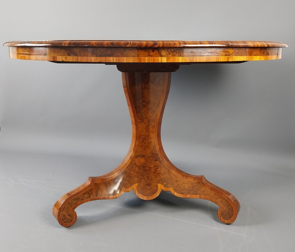 A nineteenth-century circular burr walnut centre table featuring a highly figured top, crossbanded border, and a carved pedestal on four scroll feet. View V