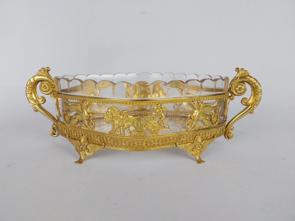 An oval cut glass bowl with a scalloped rim housed within an ornate gilt bronze mount featuring winged sphinxes, lion chariots, and scrolled handles.