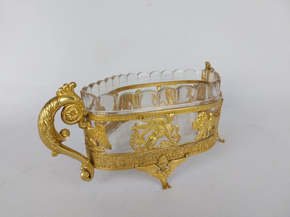 An oval cut glass bowl with a scalloped rim housed within an ornate gilt bronze mount featuring winged sphinxes, lion chariots, and scrolled handles. View X