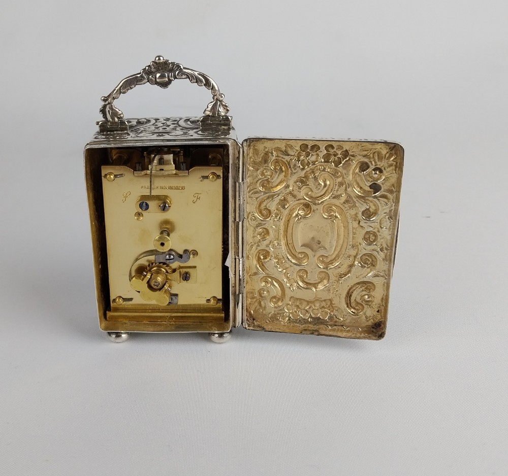 A rectangular miniature silver carriage clock featuring floral relief chasing, a curved carrying handle, ball feet, and a white enamel dial with Roman numerals. View VIII