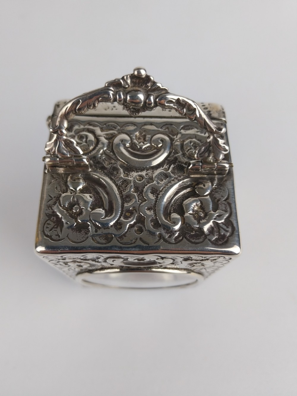 A rectangular miniature silver carriage clock featuring floral relief chasing, a curved carrying handle, ball feet, and a white enamel dial with Roman numerals. View XIII